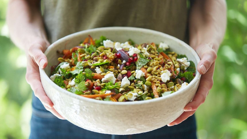 Two hands hold a serving bowl of grain salad for a celebration, with freekeh, pistachio, goat cheese, roasted carrots and herbs.