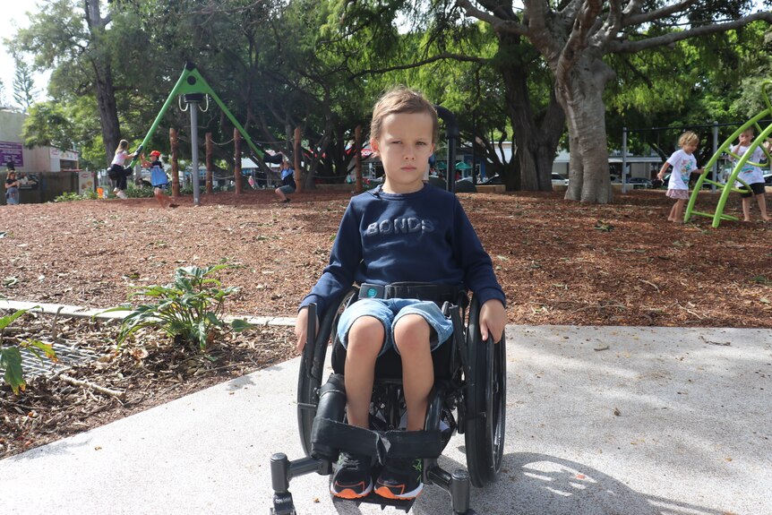 A young boy in navy sitting in his wheelchair on the concrete edge of Brennan Park.