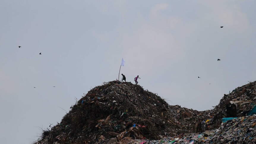 Two men walk down a large garbage mound with a white flag on top of it.