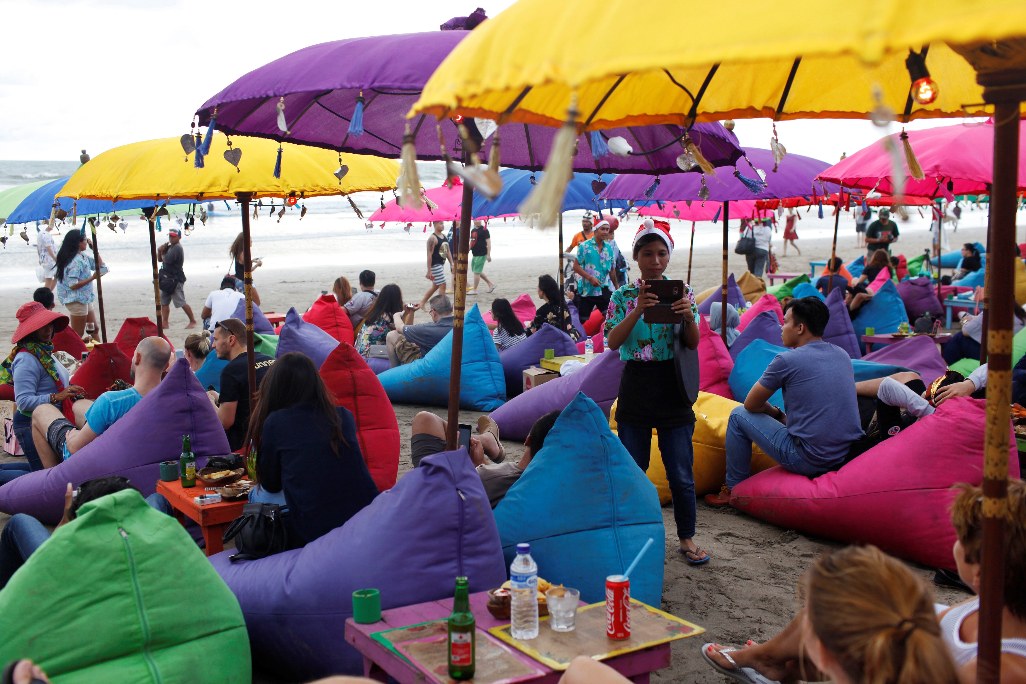 Pessoas relaxando na praia, sentadas em pufes sob um guarda-chuva e desfrutando de bebidas