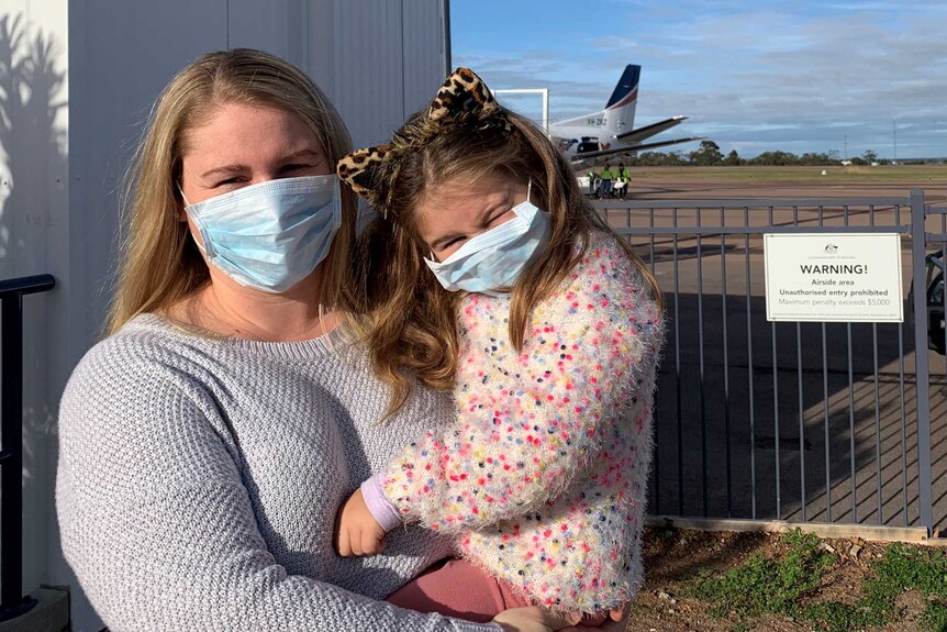 A woman holding a toddler at an airport while both wear a face mask.