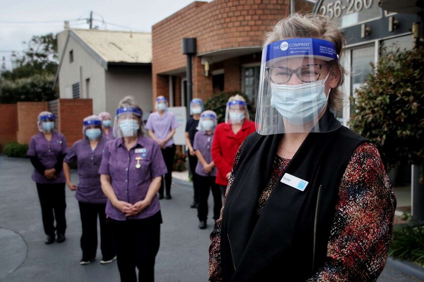 A woman with short blonde hair stands in a mask and face shield smiling. Behind her is six staff spread out in masks and shields