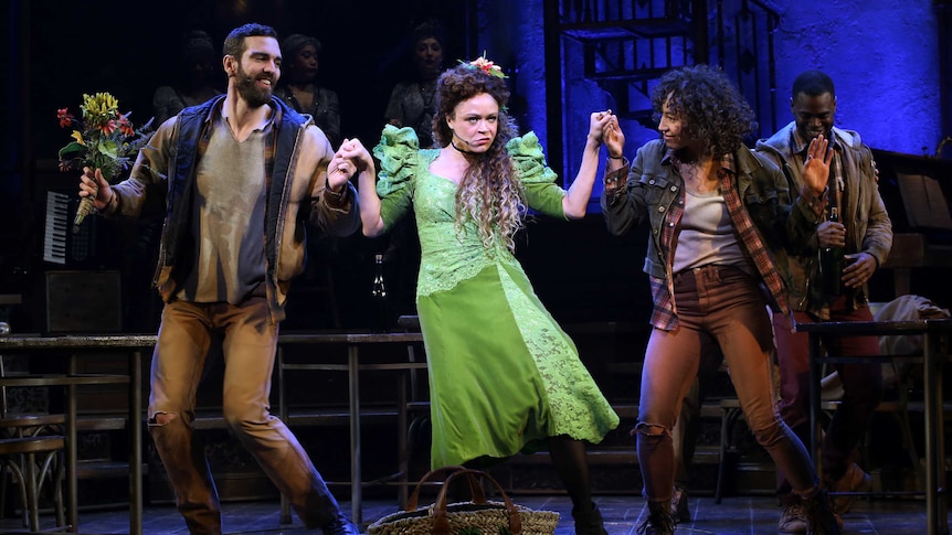A man and two women hold hands and dance on stage in the musical Hadestown.