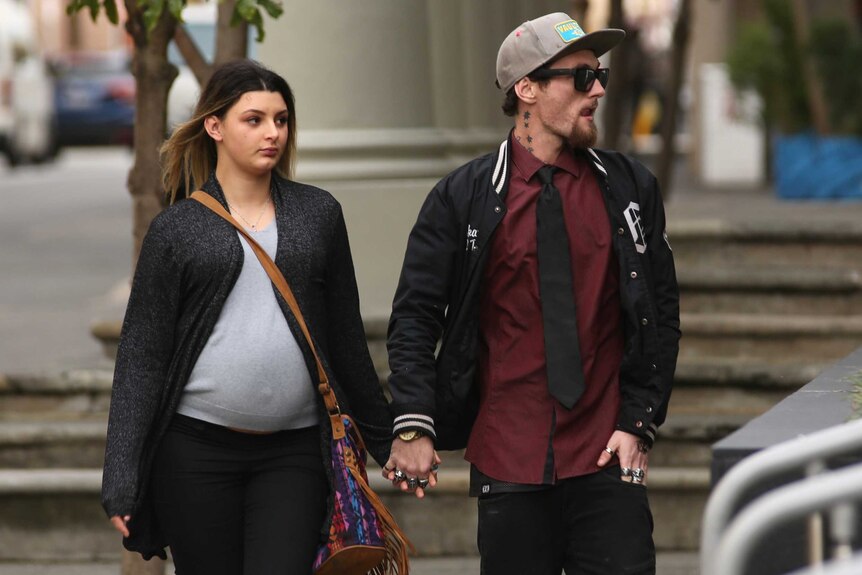 A young man in sunglasses, a baseball cap and black jacket arrives at court holding hands with a heavily pregnant woman.