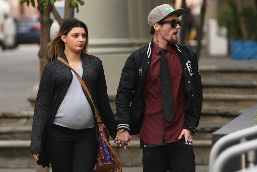 A young man in sunglasses, a baseball cap and black jacket arrives at court holding hands with a heavily pregnant woman.