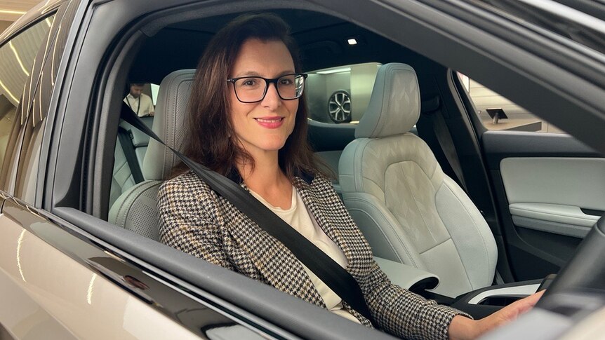 A woman with glasses sits in a car and looks out the driver's window at the camera 