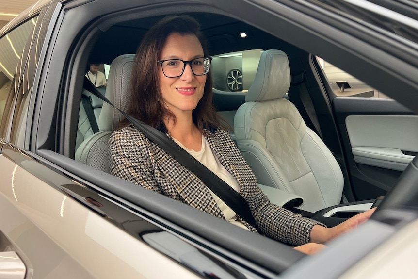 A woman with glasses sits in a car and looks out the driver's window at the camera 