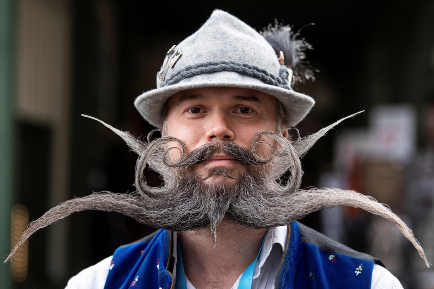 A man wears a grey hat while manicured grey beard is shaped in curly and pointed designs 