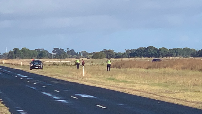 Three figures in a field next to a country road, a police car to the left, a car in a field on the right