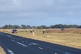 Three figures in a field next to a country road, a police car to the left, a car in a field on the right