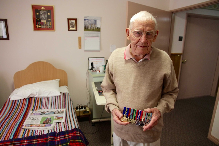 A man with white hair standing in a bedroom holding a row of medals.