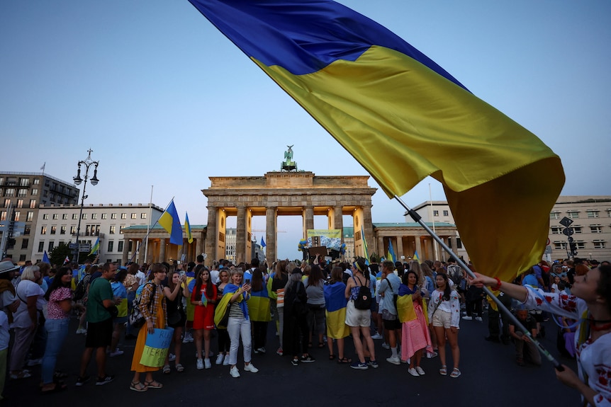 Женщина машет украинским флагом, а у Бранденбургских ворот стоит большая толпа людей.