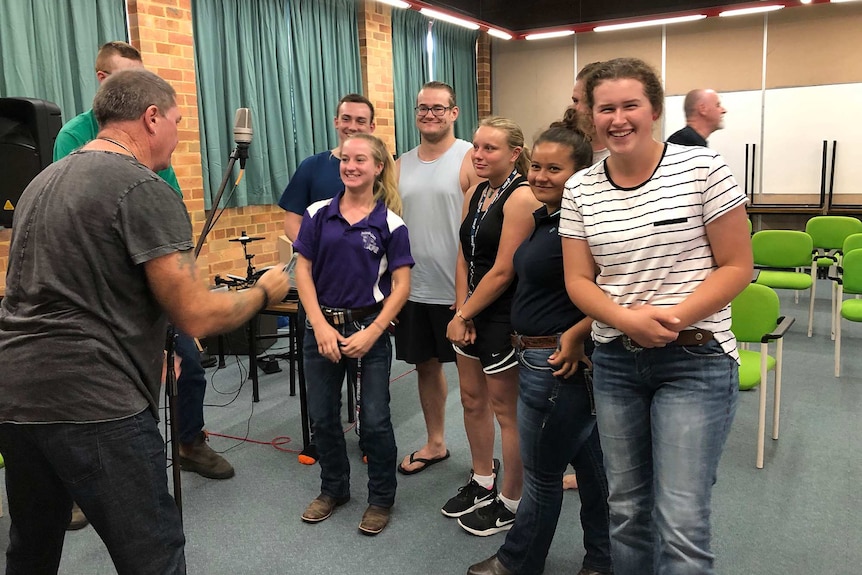 Seven students are led in singing around a microphone by a man standing in front of them.