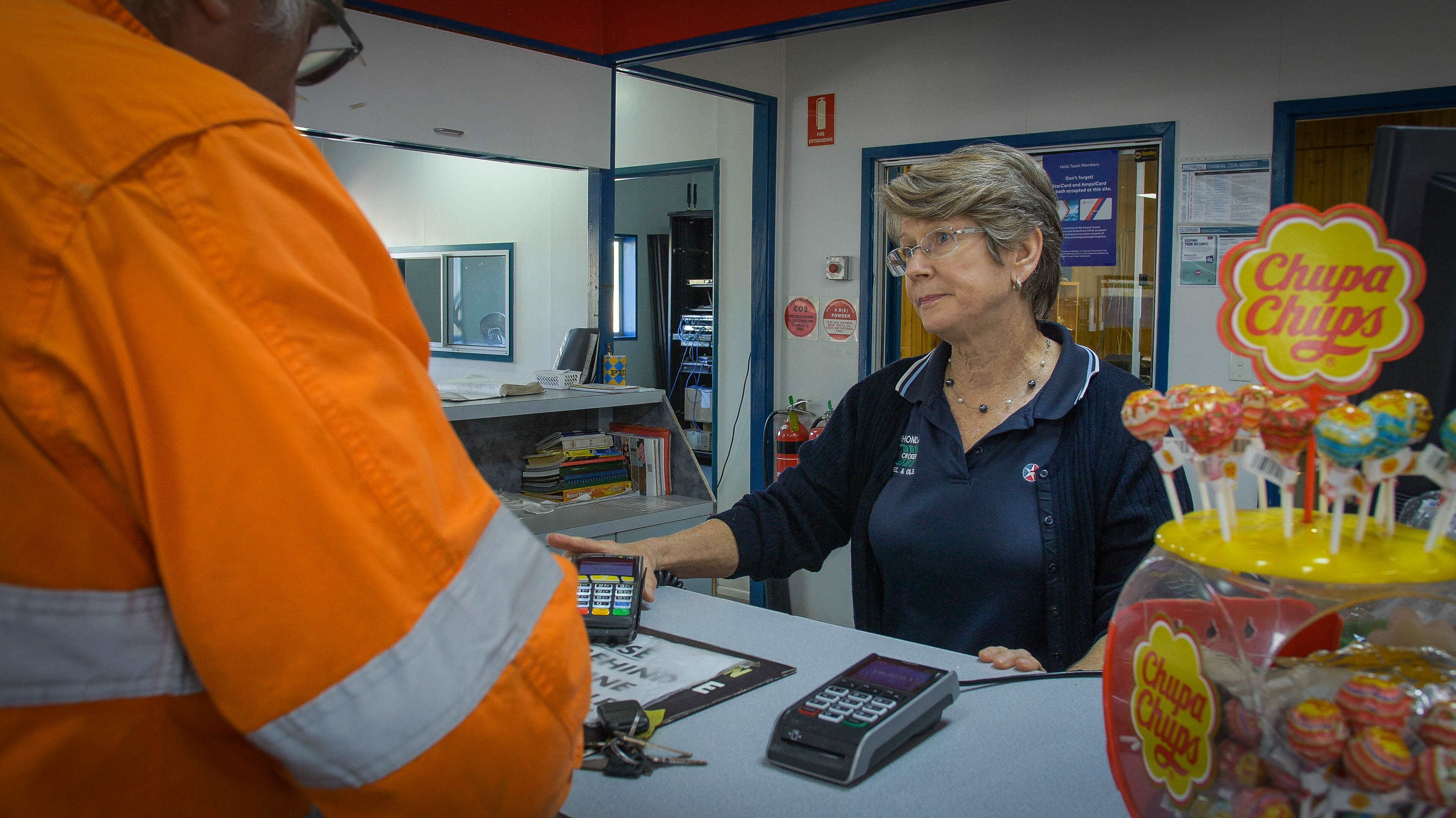 A woman stands behind a counter holding an eftpos machine, serving a man in orange high vis.
