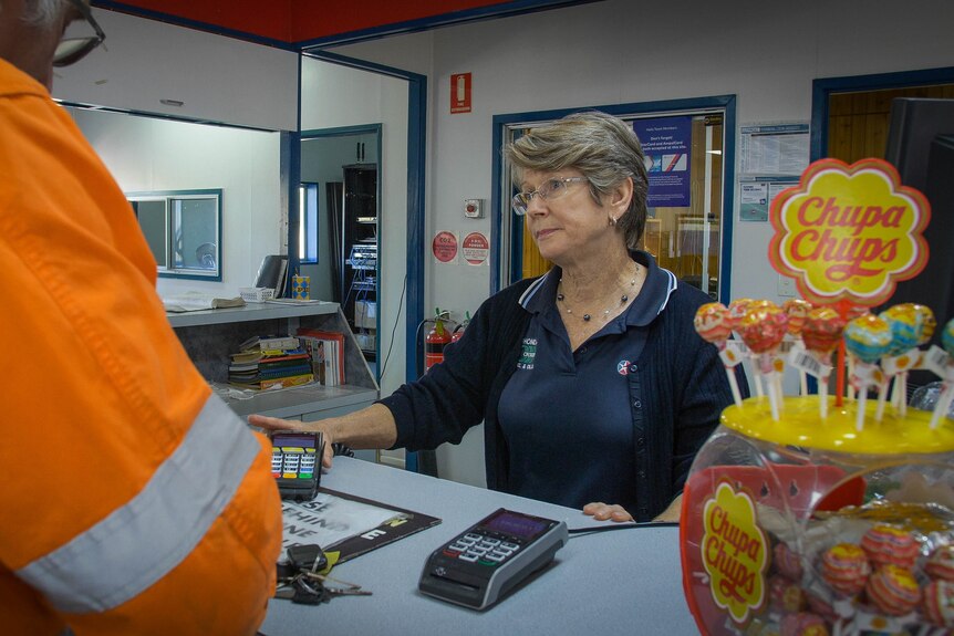 A woman stands behind a counter holding an eftpos machine, serving a man in orange high vis.