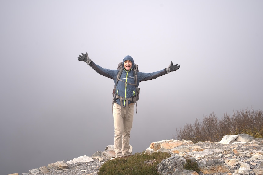 Man dressed in warm, hiking clothes standing on a rocky edge, arms out wide on a foggy day. 