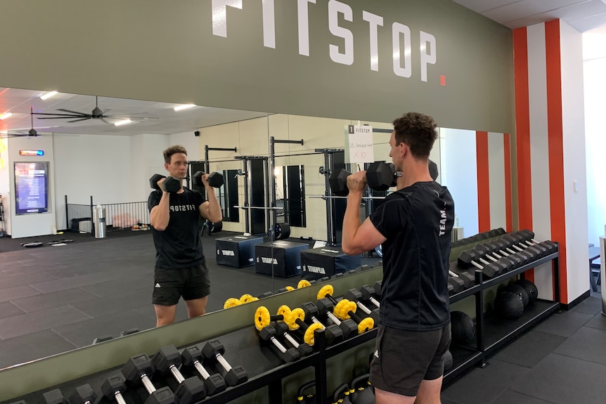 Heath Lawley stands in front of mirror, holding two dumbbells at his shoulders