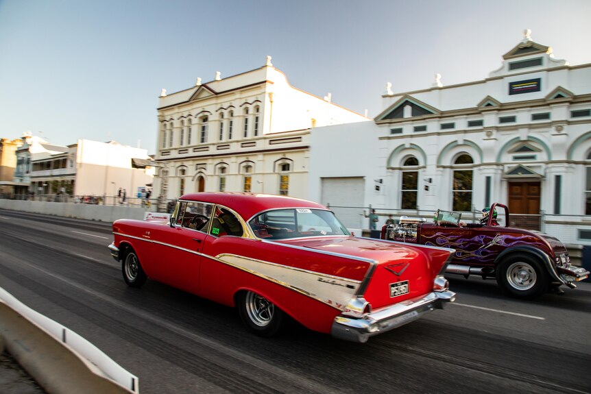 Two red vintage cars drag racing on a street, historic buildings in the background