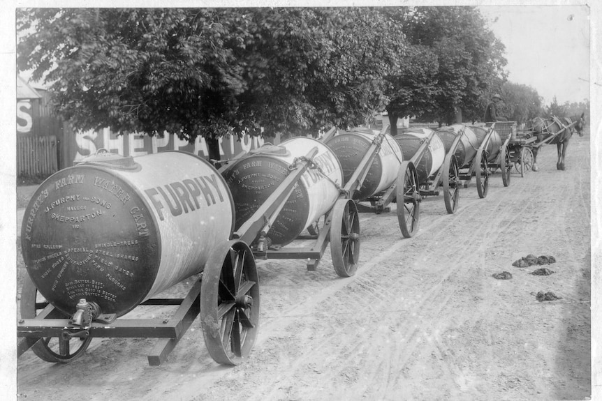 a black and white photo of a horse towing about 7 furphy water cants behind it 