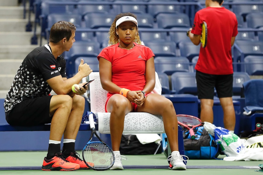 A female tennis player sits, slumped in her seat with her male coach talking to her.