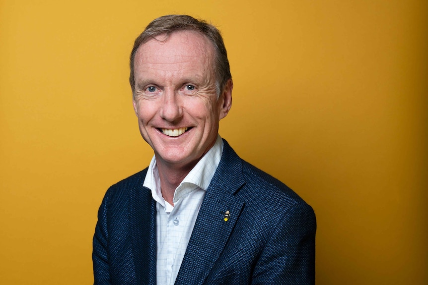 A headshot of a smiling man wearing a blue suit jacket and button up shirt on an orange background