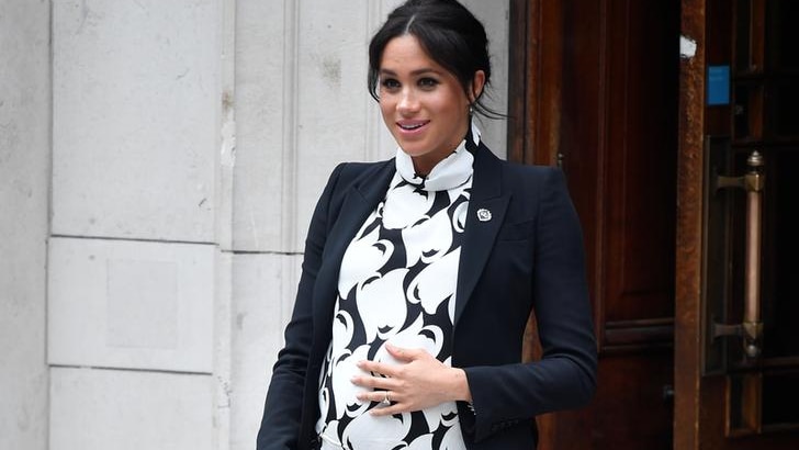 Meghan wears a black and white dress and has a hand on her bump as she exits a university building with a clutch in hand.