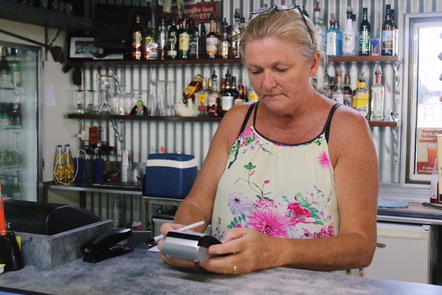 She stands at the cash register, with lots of bottles on a shelf behind her