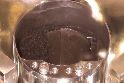 A metal cylinder displays a small load of dark material that looks a mix of pebbles and top soil.