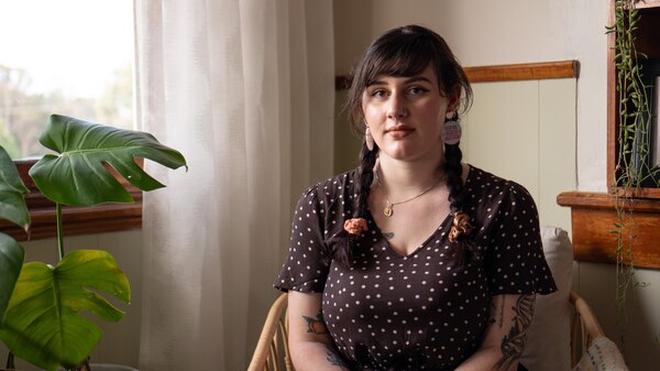 A young woman looks into the camera unsmiling, she is sat on a rattan chair next to a pot plant.