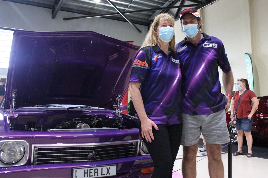 A woman and man stand in purple shirts wearing masks, in front of a purple car with the hood raised.