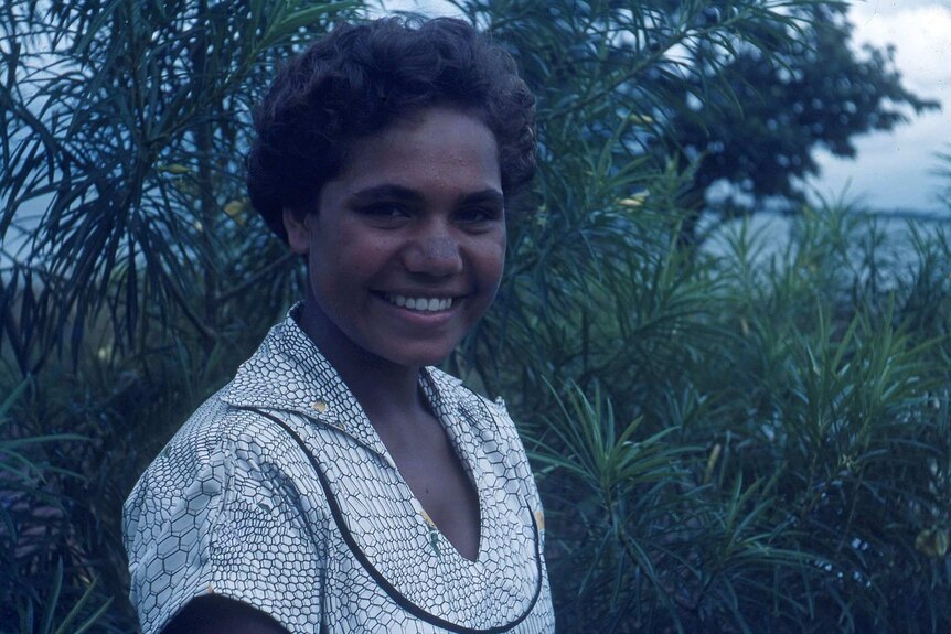 A young and smiling Indigenous woman in a white and patterned dress in front of a verdant backdrop.