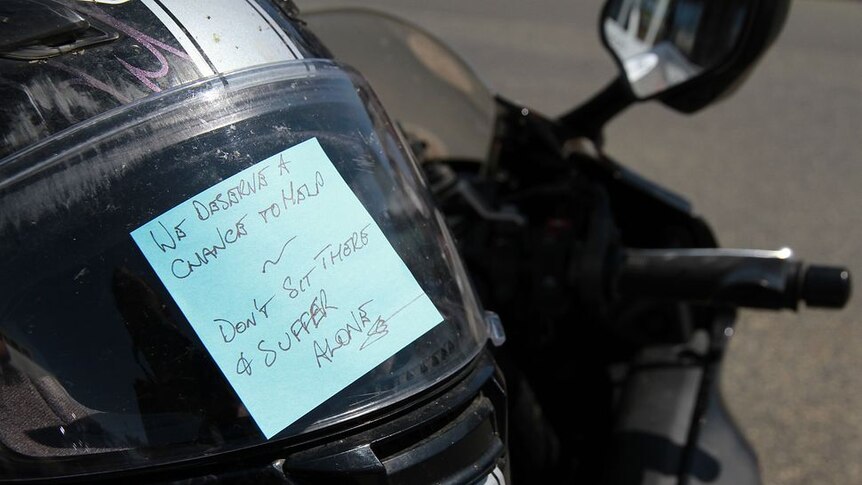 Motorbike windshield with a post-it note reading 'we deserve a chance to help: don't sit there and suffer' stuck to it
