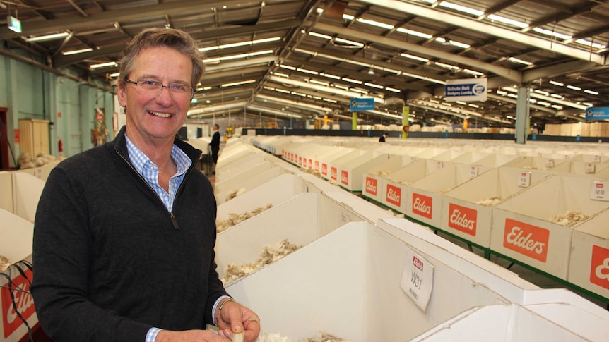 Man in charcoal jumper surrounded by boxes on Merino wool in a big warehouse. Man is smiling at the camera holding some wool.