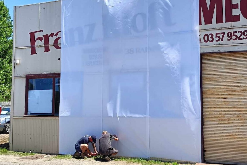 Two people setting up a white backdrop sheet for a projected display over the front of a building.