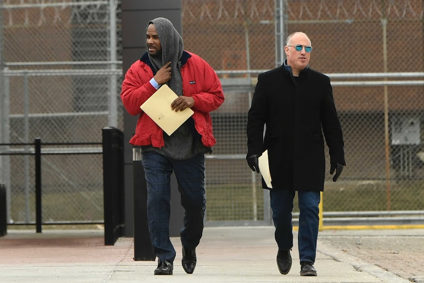 Singer R Kelly walking with a barbed wire fence hind him and a man next to him