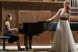 A woman in a black dress sites at a grand piano, accompanying a woman singer wearing a long white gown.
