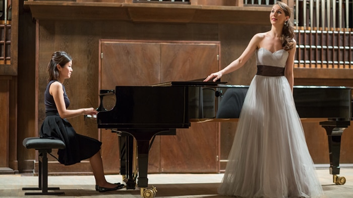 A woman in a black dress sites at a grand piano, accompanying a woman singer wearing a long white gown.