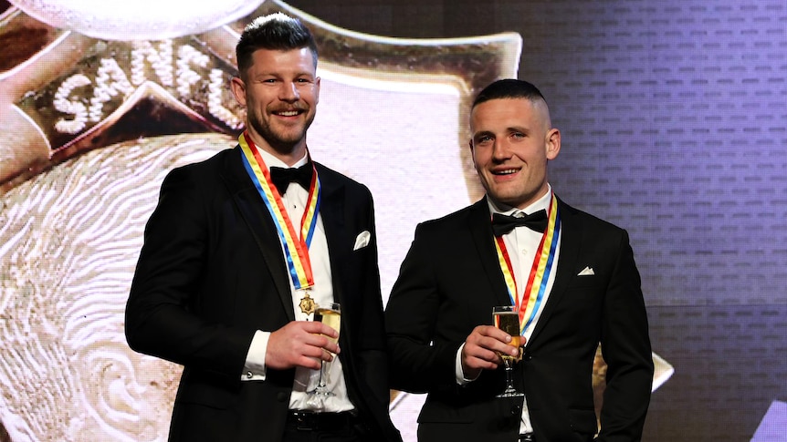Two men in tuxedos with medals around their neck smile holding sparkling wine.