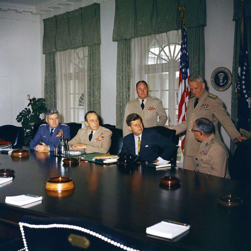 JFK sitting at a large wooden table, with 3 military leaders seated beside him and 2 standing behind him.