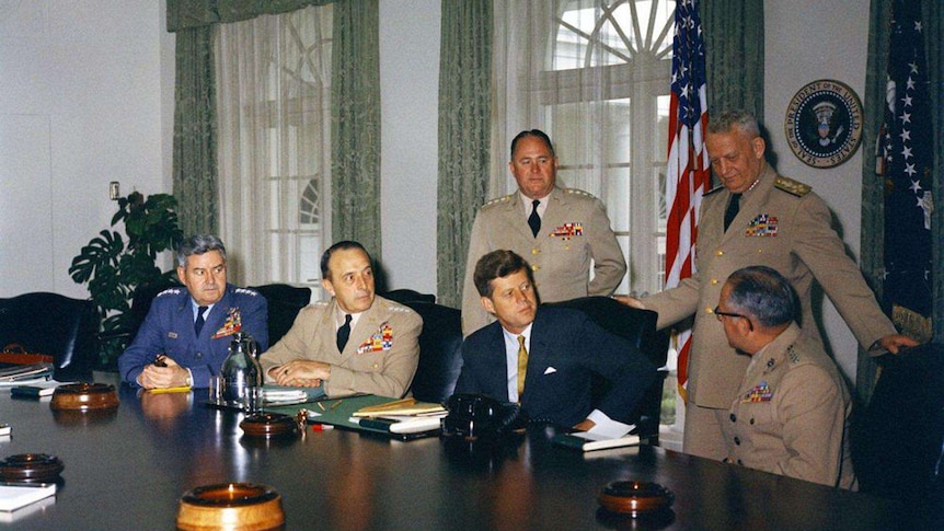 JFK sitting at a large wooden table, with 3 military leaders seated beside him and 2 standing behind him.
