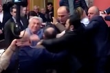 A group of men in suits scramble, blurred a bit by the movement. One is reaching across another.