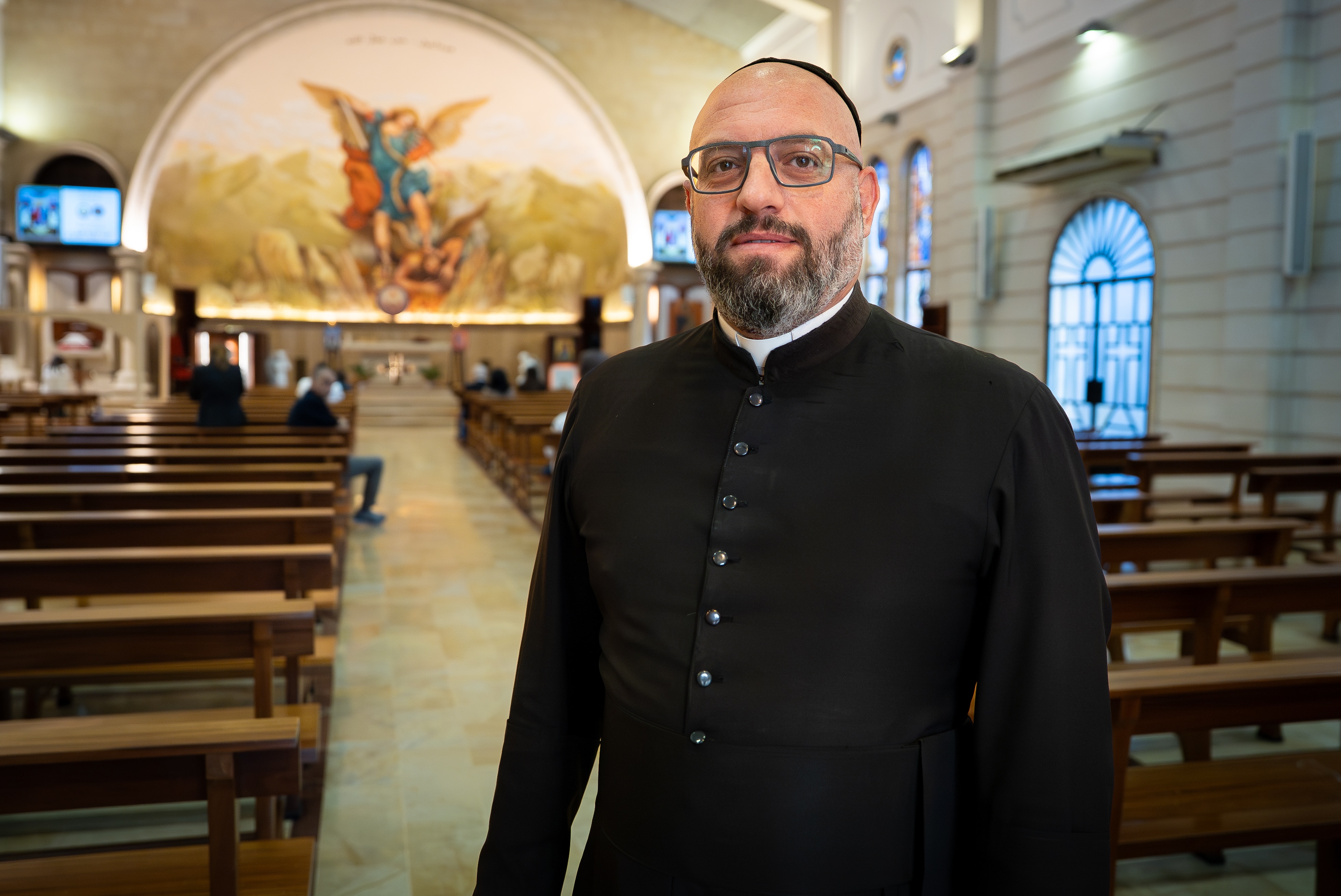 A priest with a beard wearing all black standing in a church with the benches behind him