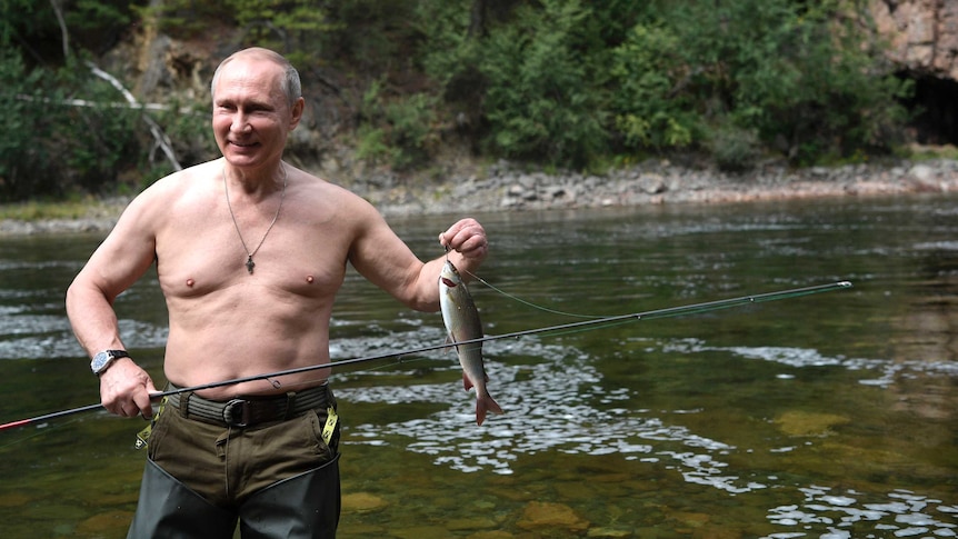 A shirtless Vladimir Putin stands on a river bank, holding up a fish still attached to his hook.