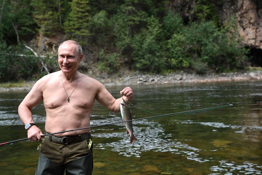 A shirtless Vladimir Putin stands on a river bank, holding up a fish still attached to his hook.