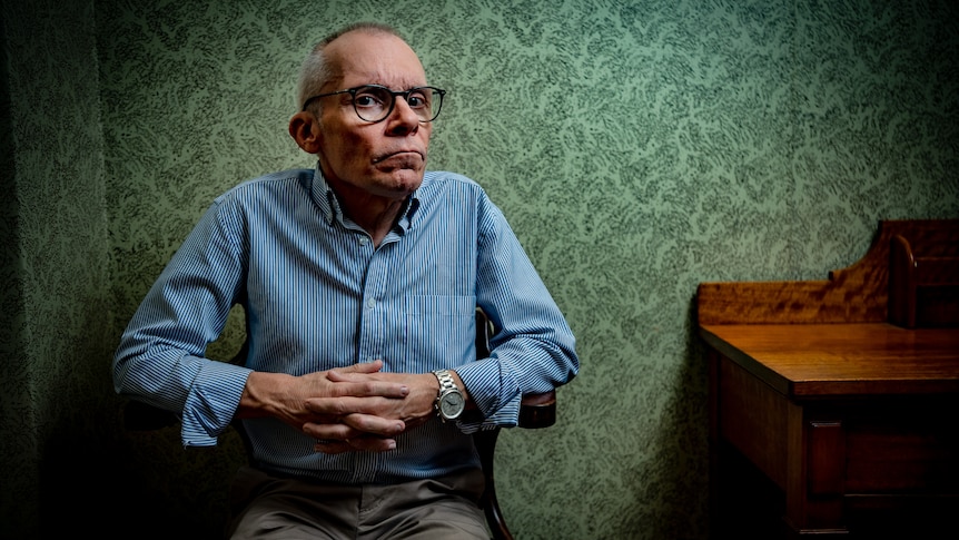 Elderly man in a blue shirt sitting in a chair with a green wallpaper behind him.