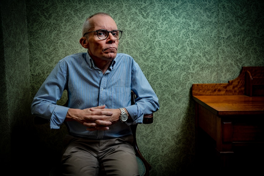 Elderly man in a blue shirt sitting in a chair with a green wallpaper behind him.