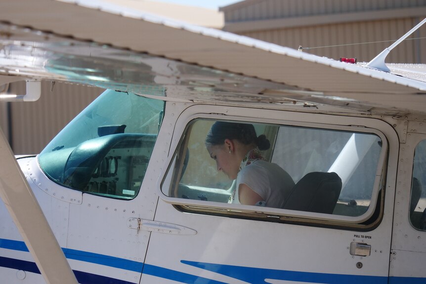 A young woman in a white shirt sits in the pilot seat of a small white airplane, looking down at what's in front of her.