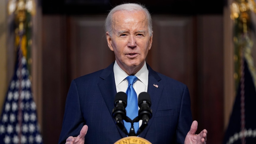 An image of Joe Biden speaking in front of a microphone. He is wearing a dark blue suit and a blue tie.