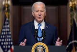 An image of Joe Biden speaking in front of a microphone. He is wearing a dark blue suit and a blue tie.