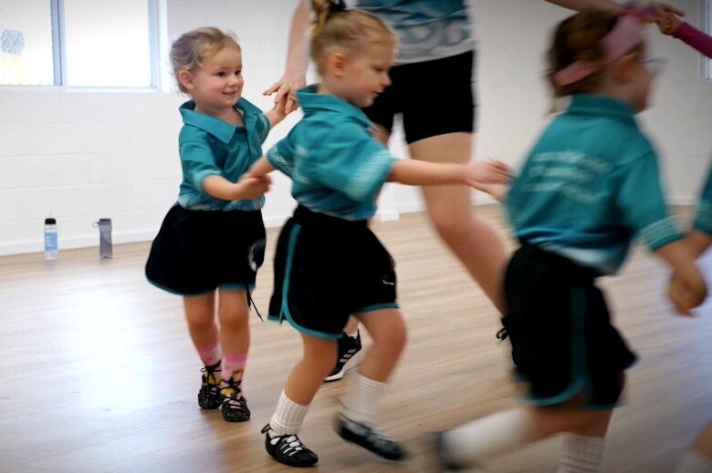 Children in green tops and black shorts uniforms holding hands and dancing in a circle in a dancehall